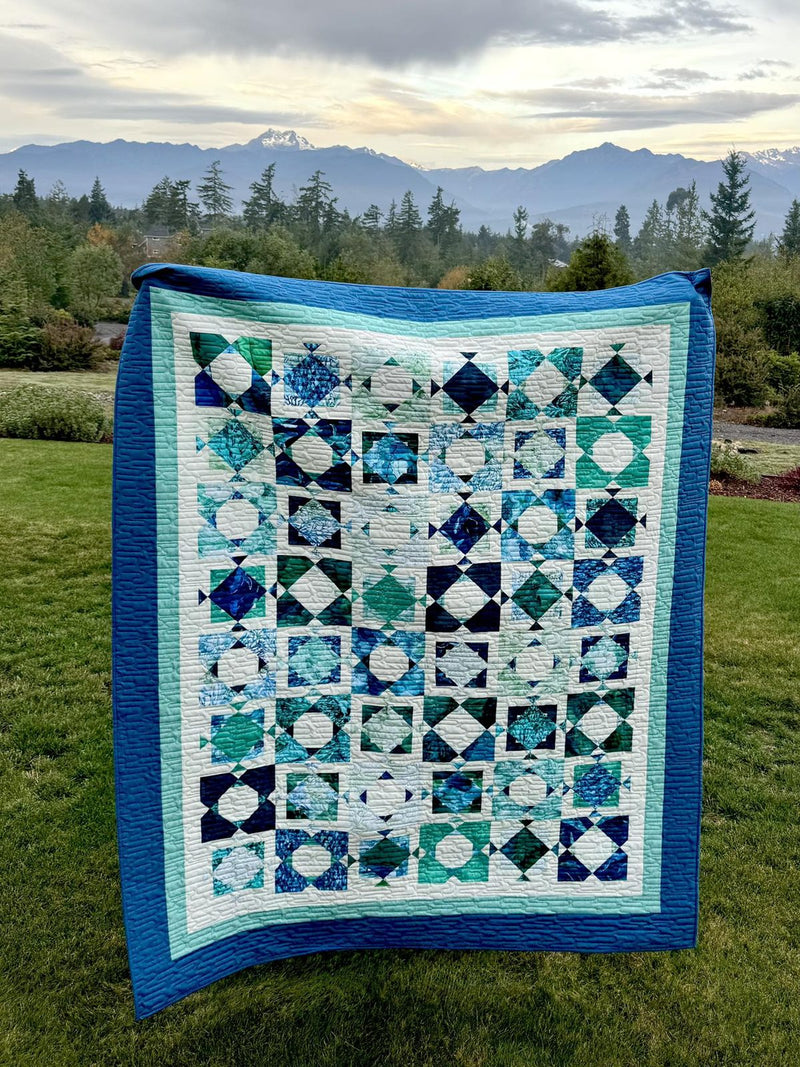 A finished blue and white quilt held up outdoors in front of mountains
