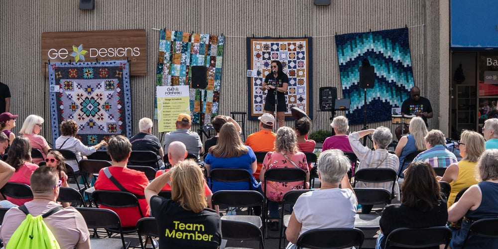 Gudrun Erla speaking on the GE FamFest stage outside the GE Designs building, with a crowd of quilters seated in front of her